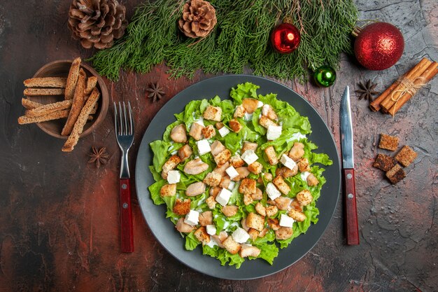 Vue de dessus salade césar sur plaque ovale fourchette couteau bol avec croûte séchée arbre de Noël jouets bâtons de cannelle sur fond rouge foncé