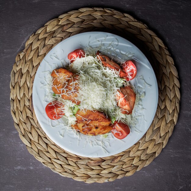 Vue de dessus salade César avec poulet, tomate, laitue, olives, craquelins, parmesan dans une assiette blanche sur un support en osier