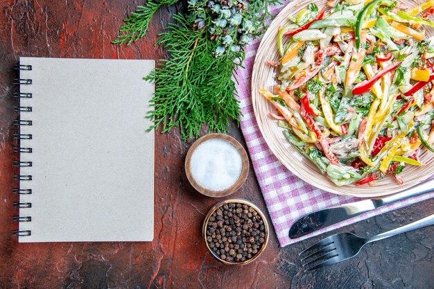 Vue de dessus salade de légumes sur plaque sur nappe fourchette et couteau sel et poivre noir un cahier sur table rouge foncé