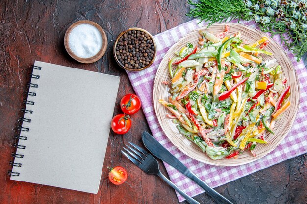 Vue de dessus salade de légumes sur plaque sur nappe fourchette et couteau sel et poivre noir tomates un cahier sur table rouge foncé