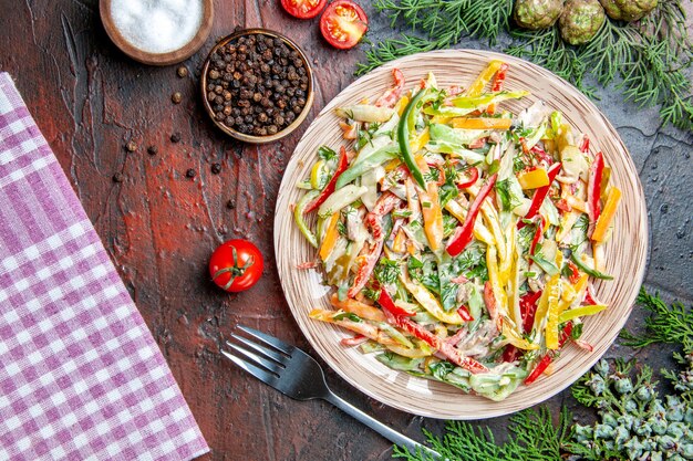 Vue de dessus salade de légumes sur plaque nappe sel fourchette et poivre noir tomates branches de pin sur table rouge foncé