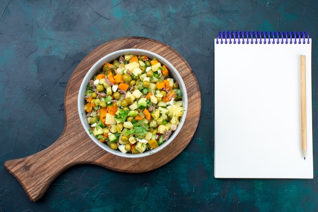 Vue de dessus salade de légumes en tranches poivrée de tranches de poulet à l'intérieur de la plaque avec bloc-notes sur le bureau bleu foncé