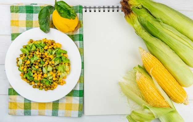 Vue de dessus de la salade de maïs sur tissu et citron avec épis de maïs et bloc-notes sur la surface en bois