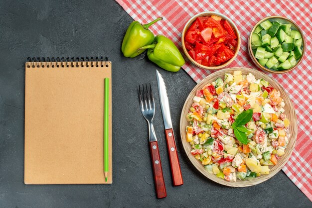 Vue de dessus de la salade végétarienne avec serviette rouge en dessous et couverts de légumes et bloc-notes sur le côté sur fond gris foncé