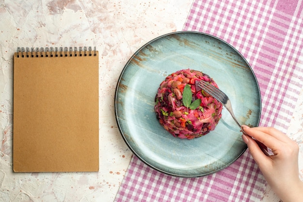 Vue de dessus de la salade de vinaigrette sur une assiette ovale sur une nappe à carreaux blancs et violets une fourchette dans un bloc-notes à main de femme sur une table gris clair