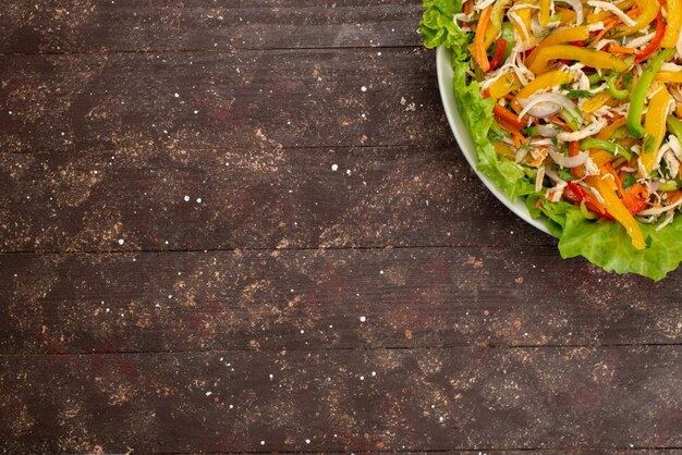 Vue de dessus savoureuse salade de légumes avec des légumes tranchés et salade verte à l'intérieur d'une assiette ronde sur brun, repas de salade de légumes