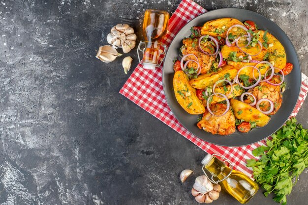 Vue de dessus d'un savoureux repas de poulet avec des pommes de terre et des légumes verts sur une serviette rouge dépouillée des bouteilles d'huile tombées sur le côté gauche sur une table de couleur sombre