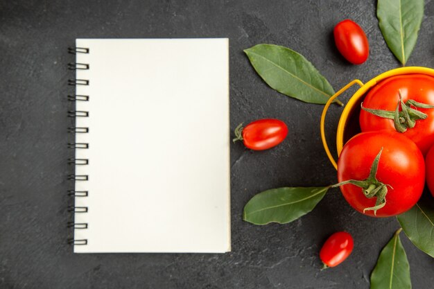 Vue de dessus un seau avec des tomates autour des tomates cerises et des feuilles de laurier et un ordinateur portable sur fond sombre