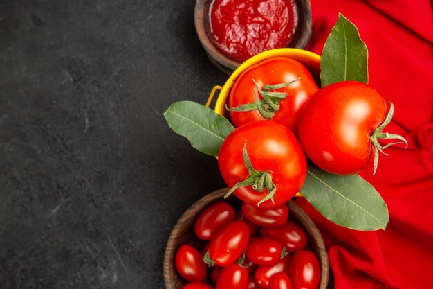 Vue de dessus un seau avec des tomates et des bols de feuilles de laurier avec des tomates cerises et du ketchup et une serviette rouge sur fond sombre