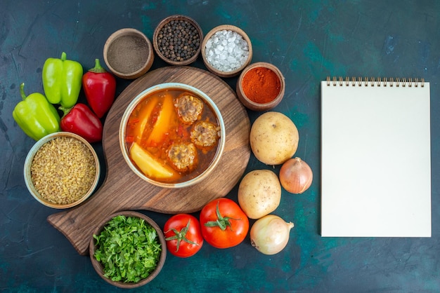 Vue de dessus de la soupe de boulettes de viande avec des pommes de terre tranchées à l'intérieur et des légumes frais sur un bureau bleu foncé