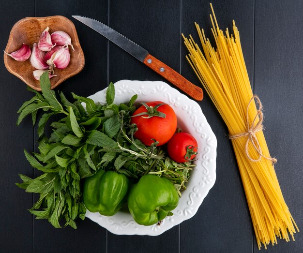 Vue de dessus des spaghettis crus avec tomates menthe et poivron dans une assiette avec un couteau et de l'ail sur une surface noire