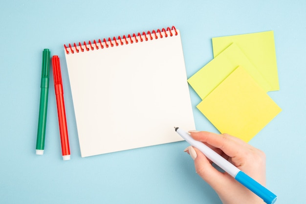 Vue de dessus stylo à la main papiers collants jaunes marqueurs bloc-notes sur bleu