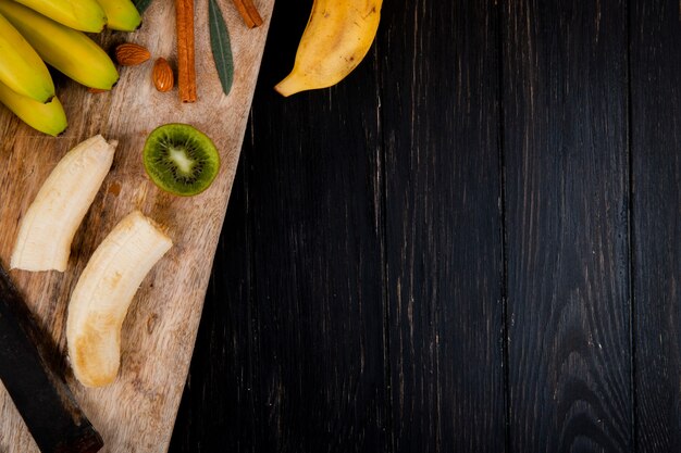 Vue de dessus d'un tas de banane aux amandes, bâtons de cannelle et vieux couteau de cuisine sur une planche à découper en bois sur fond noir avec copie espace