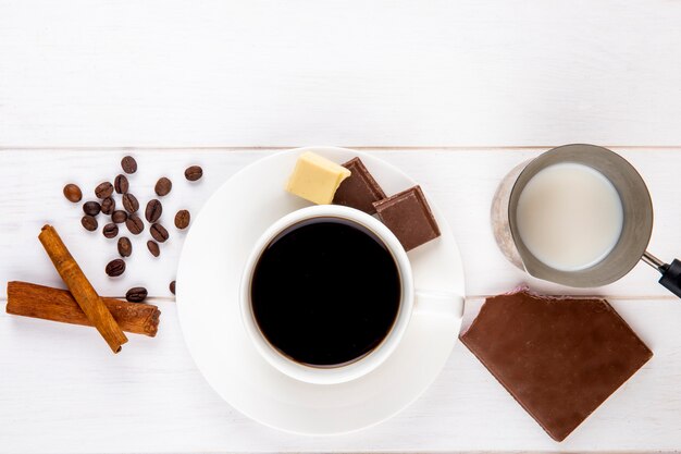 Vue de dessus d'une tasse de café avec des bâtons de cannelle barre de chocolat et grains de café éparpillés sur fond de bois blanc