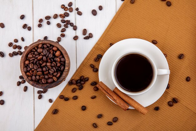 Vue de dessus d'une tasse de café sur un chiffon avec des bâtons de cannelle avec des grains de café sur un bol en bois sur fond blanc