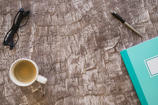 Une vue de dessus d&#39;une tasse de café; stylo; cahier et lunettes sur fond texturé