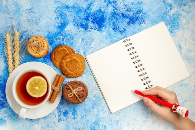 Vue de dessus tasse de stylo bloc-notes de biscuits au thé dans une main féminine sur une table bleue