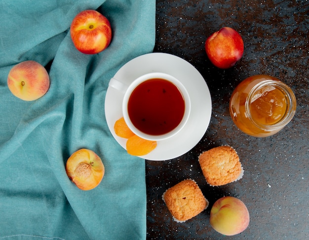 Vue De Dessus D'une Tasse De Thé Avec Des Abricots Secs Et Des Pêches Mûres Fraîches Sur Tissu Bleu Et Des Muffins Avec Un Bocal En Verre De Confiture De Pêches Sur Fond Noir