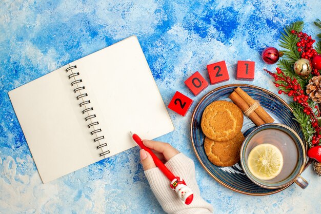 Vue de dessus tasse de thé aromatisé par des bâtons de cannelle citron biscuits sur soucoupe stylo bloc-notes dans la main féminine sur la table bleue
