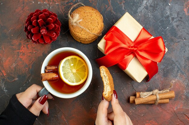 Vue de dessus tasse de thé aromatisée au citron et à la cannelle et biscuit dans la main féminine cadeau de Noël pomme de pin rouge bâtons de cannelle biscuits sur table rouge foncé