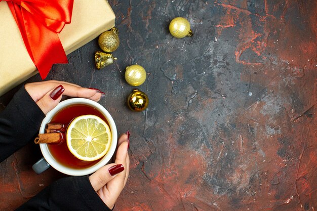 Vue de dessus tasse de thé aromatisée au citron et à la cannelle dans des boules de Noël dorées à la main féminine sur une table rouge foncé