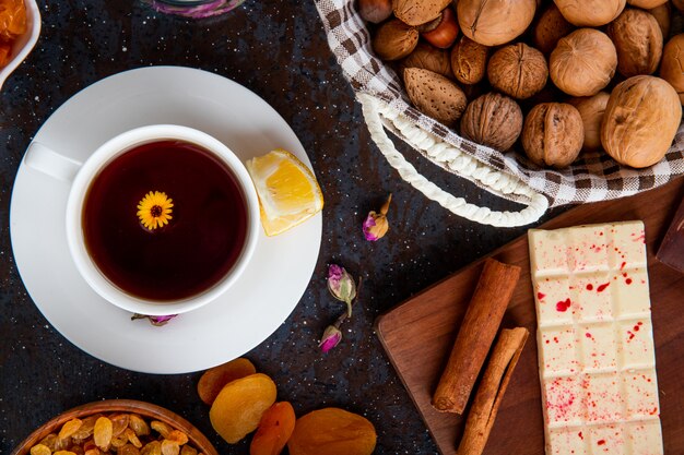 Vue de dessus d'une tasse de thé au citron, avec barre de chocolat blanc, fruits secs et noix sur fond noir