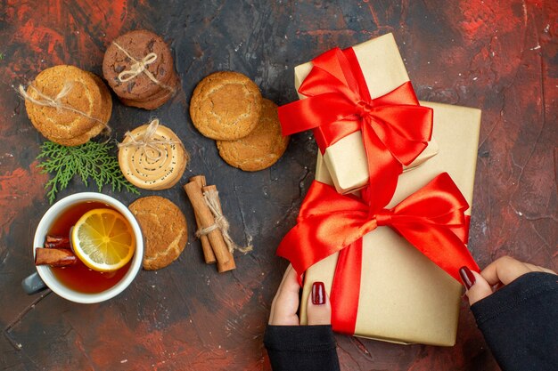 Vue de dessus tasse de thé bâtons de cannelle différents biscuits attachés avec une corde présents dans la main féminine sur une table rouge foncé