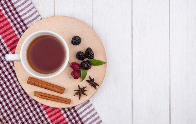 Vue de dessus de la tasse de thé à la cannelle, framboises et mûres sur un support sur une surface blanche