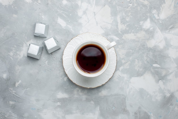 Vue de dessus de la tasse de thé chaud à l'intérieur tasse blanche sur plaque de verre avec des bonbons en argent sur un bureau léger, boire du thé biscuit au chocolat sucré