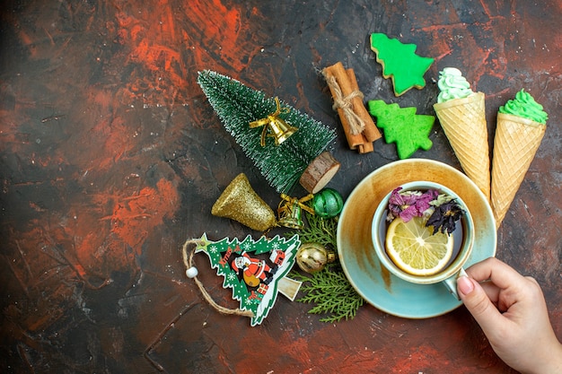 Photo gratuite vue de dessus tasse de thé dans les glaces à la main féminine bâtons de cannelle biscuits d'arbre de noël ornements de noël sur table rouge foncé