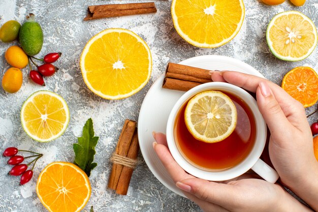 Vue de dessus une tasse de thé dans les mains de la femme coupe des bâtons de cannelle citrons sur fond gris