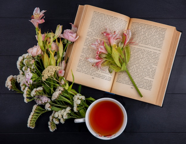 Vue de dessus de la tasse de thé avec des fleurs rose clair et un livre ouvert sur une surface noire