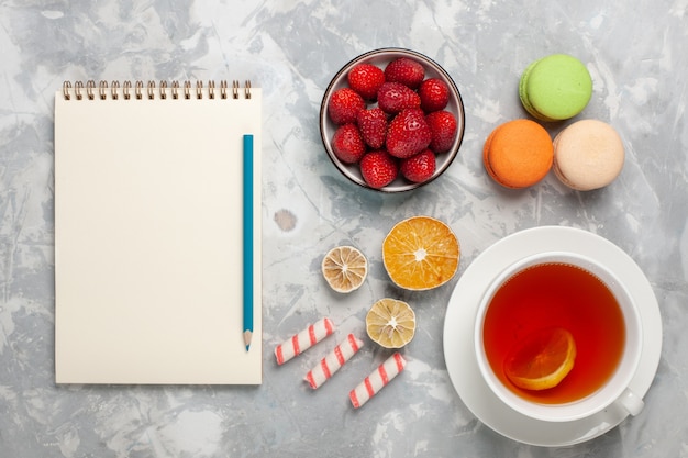 Vue de dessus tasse de thé avec des fraises rouges fraîches et des macarons français sur un bureau blanc