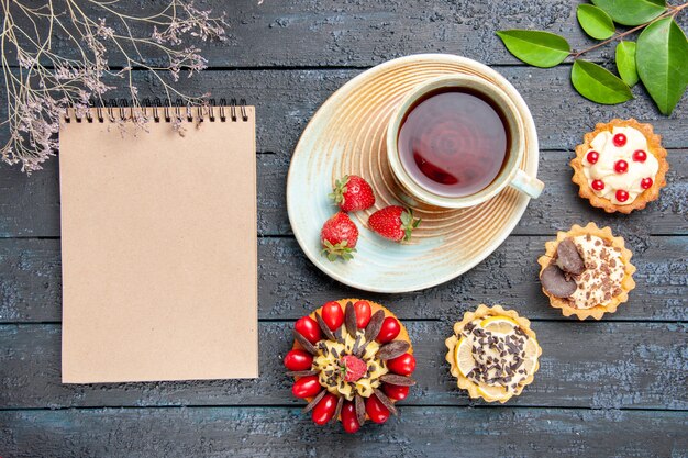 Photo gratuite vue de dessus une tasse de thé et de fraises sur soucoupe oranges séchées tartelettes laisse gâteau aux baies et un cahier sur la table en bois sombre
