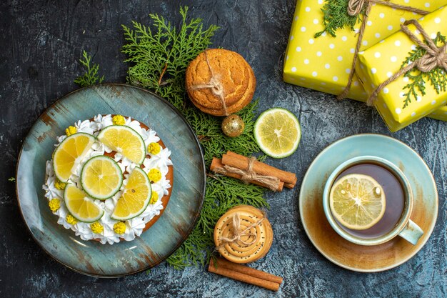 Vue de dessus d'une tasse de thé noir délicieux gâteau crémeux et branches de sapin citron cannelle limes coffrets cadeaux jaunes sur fond sombre