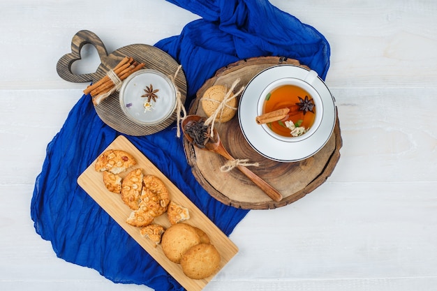 Vue de dessus de la tasse de thé sur une planche de bois avec des biscuits et de la cannelle sur des planches à découper, foulard bleu sur une surface blanche