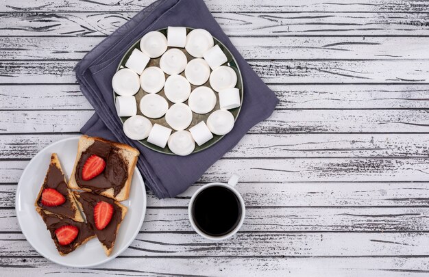 Vue de dessus des toasts de petit déjeuner avec chocolat et fraise, guimauve et café sur une surface en bois blanche horizontale