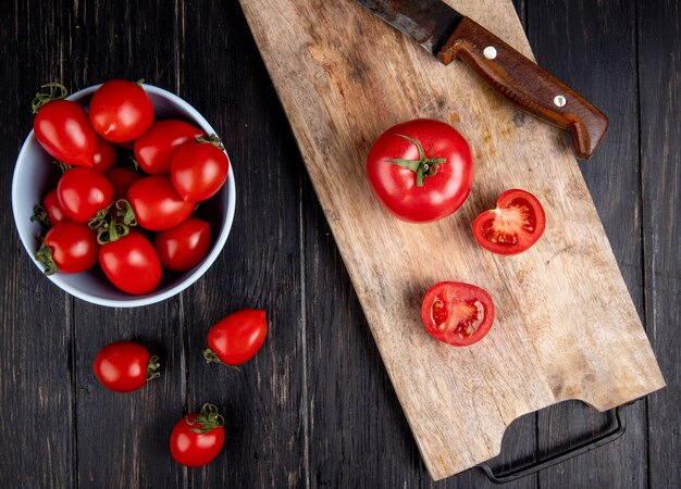 Vue de dessus des tomates coupées et entières et couteau sur une planche à découper avec d'autres dans un bol sur une surface en bois