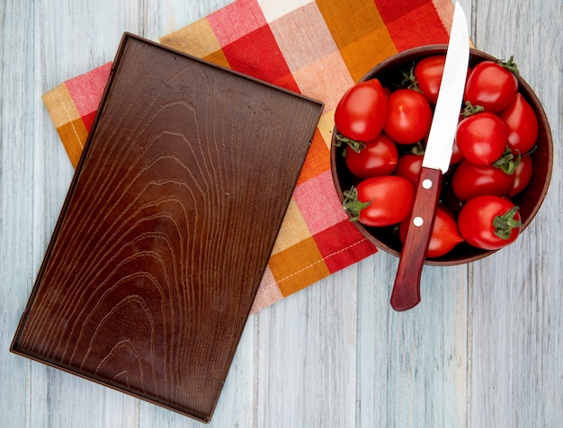 Vue de dessus des tomates avec un couteau dans un bol et un plateau vide sur un chiffon sur une surface en bois