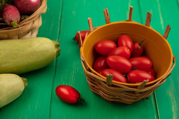 Vue de dessus de tomates prunes rouges fraîches sur un seau avec des radis sur un seau avec des courgettes isolé sur un mur en bois vert