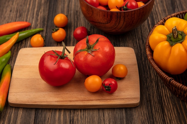 Vue de dessus des tomates rouges de grande taille sur une planche de cuisine en bois avec des tomates cerises sur un bol en bois et des poivrons sur un seau sur une surface en bois