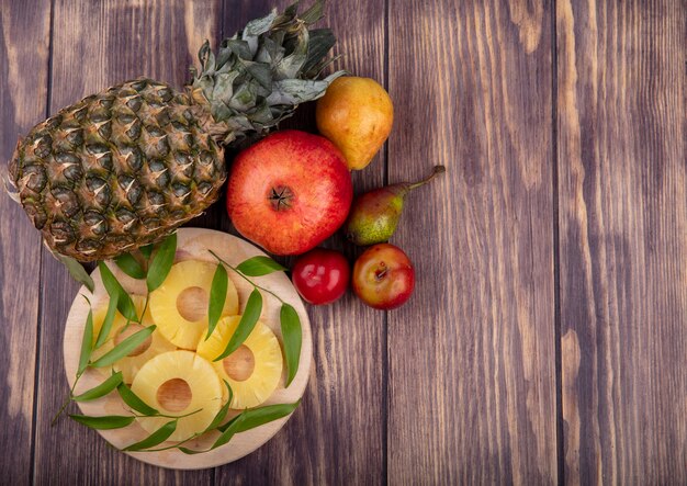 Vue de dessus des tranches d'ananas avec des feuilles sur une planche à découper et ananas grenade pêche prune sur une surface en bois