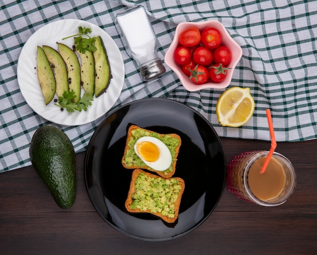 Vue de dessus des tranches d'avocat sur plaque blanche avec tomates avocat frais tranches de pain toast lemonnd avec pulpes d'avocat et oeuf sur nappe à carreaux et bois