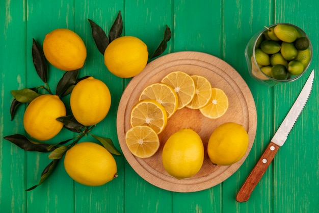 Vue de dessus des tranches de citrons sur une planche de cuisine en bois avec un couteau avec des kinkans sur un verre avec des citrons isolé sur un mur en bois vert