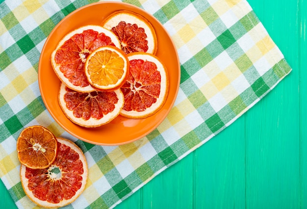 Vue de dessus des tranches d'orange et de pamplemousse séchées dans une assiette sur une nappe à carreaux sur fond de bois vert avec copie espace