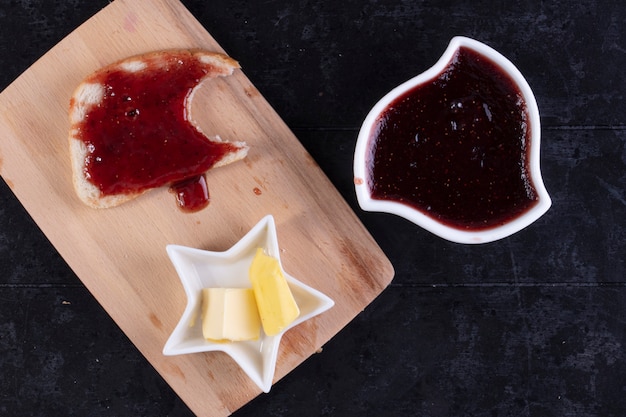 Vue de dessus des tranches de pain avec de la confiture sur tableau noir avec du beurre et de la confiture dans une soucoupe sur fond noir