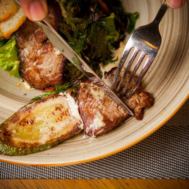 Vue de dessus de la viande frite avec des courgettes frites et une fourchette et un couteau et des légumes verts dans une assiette ronde