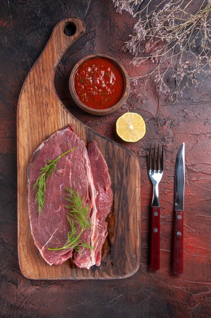 Vue de dessus de la viande rouge sur une planche à découper en bois et du ketchup dans un petit bol fourchette et couteau sur fond sombre