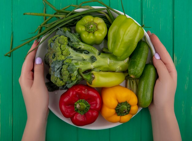 Vue de dessus woman holding brocoli avec des concombres poivrons colorés et oignons verts sur une assiette sur un fond vert