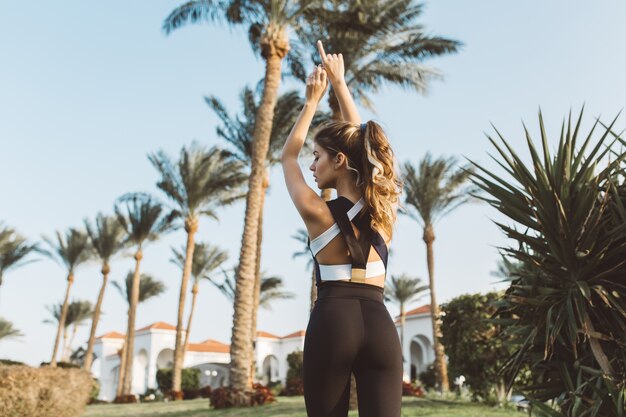 Vue de dos incroyable jolie femme en vêtements de sport strecthing dans la ville tropicale. Matin ensoleillé, palmiers, vraies émotions, mode de vie sain, entraînement, modèle à la mode